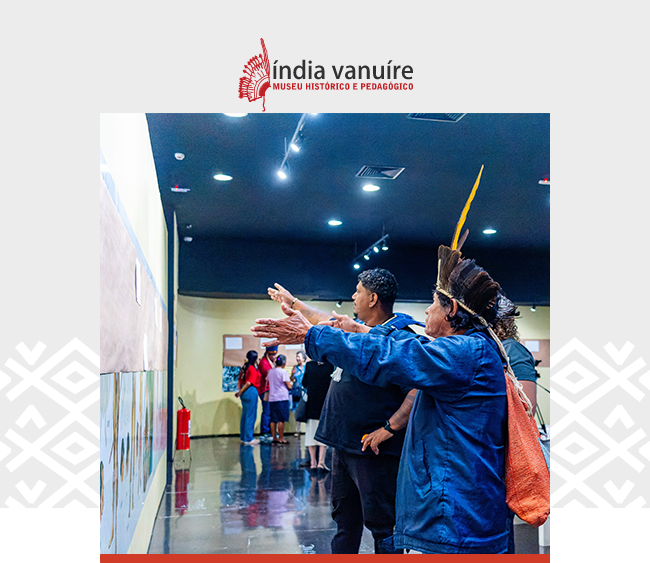 Na parte superior, o logotipo do Museu Índia Vanuíre com a silhueta estilizada de um adorno de cabeça indígena, seguido do texto “Índia Vanuíre” em letras pretas e vermelhas, e da inscrição “Museu Histórico e Pedagógico”. Abaixo, uma fotografia mostra um ambiente interno iluminado, com piso brilhante e teto escuro pontuado por luzes. Em primeiro plano, um homem indígena de perfil usa adorno de penas amarelas e jaqueta azul, estendendo os braços para frente. Ao fundo, outras pessoas observam as obras expostas na parede.