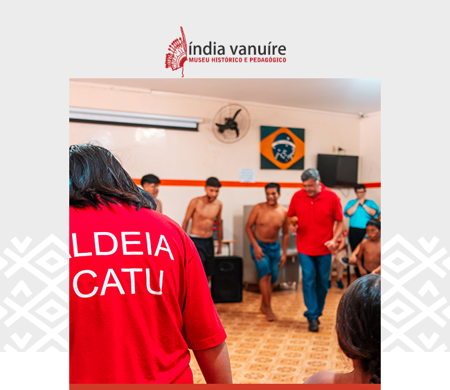 Imagem com o logotipo do Museu Índia Vanuíre centralizado na parte superior, sobre fundo claro decorado com grafismos nas laterais. Abaixo, a fotografia principal mostra um grupo reunido em um ambiente interno, durante uma atividade cultural. Em primeiro plano, aparece uma pessoa de costas vestindo camiseta vermelha com a inscrição “Aldeia Icatu”. Ao fundo, jovens — alguns sem camisa e outros com roupas leves — participam da dinâmica junto a um homem, também de camisa vermelha. A sala possui piso estampado, ventiladores na parede e bandeiras expostas, incluindo a do Brasil.
