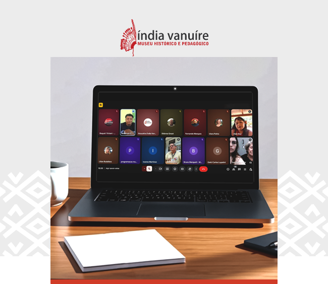 Imagem com o logotipo do Museu Índia Vanuíre centralizado na parte superior, sobre fundo claro. Abaixo, uma fotografia em destaque mostra um notebook aberto sobre uma mesa de madeira, exibindo a tela de uma reunião virtual com múltiplos participantes organizados em pequenos quadros. Algumas pessoas aparecem com câmera ligada, enquanto outras são representadas por ícones com iniciais. Sobre a mesa, à esquerda do notebook, há uma xícara branca; à frente, um bloco de anotações em branco; e, à direita, um celular apoiado próximo ao computador. O fundo é neutro, sugerindo um ambiente de trabalho ou estudo. Elementos gráficos da identidade visual do Museu Índia Vanuíre aparecem nas laterais da imagem, com uma faixa vermelha na base.