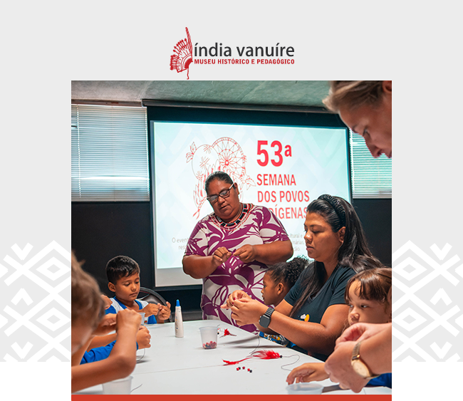 Na parte superior da imagem, o logotipo do Museu Histórico e Pedagógico Índia Vanuíre aparece centralizado sobre um fundo cinza claro, com elementos gráficos brancos nas laterais. Abaixo, a fotografia registra uma oficina educativa realizada durante a quinquagésima terceira Semana dos Povos Indígenas. No centro da imagem, uma mulher de camiseta estampada orienta um grupo de crianças e adultos sentados em torno de uma mesa. Eles manuseiam materiais coloridos, em uma atividade de artesanato. Ao fundo, um telão exibe a identidade visual do evento com os dizeres quinquagésima terceira Semana dos Povos Indígenas.