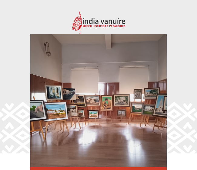 Card digital com fundo cinza-claro e o logotipo do Museu Índia Vanuíre no topo, em vermelho, com a inscrição “Museu Histórico e Pedagógico”. A imagem central mostra uma sala expositiva com piso de madeira clara, paredes revestidas em madeira até meia altura e duas janelas com persianas brancas fechadas. No espaço, há cerca de vinte pinturas emolduradas dispostas sobre cavaletes de madeira. As obras retratam paisagens, construções históricas e cenas urbanas, com cores vibrantes e estilo figurativo. A disposição dos quadros forma uma meia-lua, ocupando o centro da sala, sugerindo uma exposição temporária com foco na história e na arquitetura local. A atmosfera é acolhedora e silenciosa.
