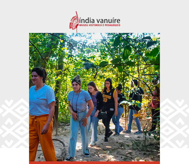 A imagem mostra um grupo de visitantes caminhando por uma trilha em meio a uma área de vegetação densa e verde, sob luz natural filtrada pelas árvores. À frente do grupo, uma mulher de camiseta branca e calça laranja conduz a caminhada, seguida por outras pessoas, majoritariamente mulheres, em clima de atenção e curiosidade. Todas caminham por um caminho de terra batida, rodeado por galhos e folhas. Na parte superior da imagem, aparece o logotipo do Museu Índia Vanuíre, com o nome da instituição em preto e vermelho e o desenho de uma mão estilizada com adorno indígena. A imagem transmite a ideia de vivência educativa em contato com a natureza, promovida pelo museu localizado em Tupã (SP).