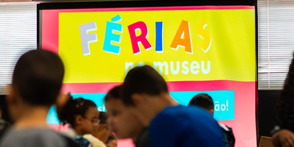 Na parte superior da imagem, uma tela exibe um cartaz com fundo amarelo e detalhes em rosa e azul. Em destaque, aparece a inscrição “FÉRIAS no museu”, com a palavra “FÉRIAS” em letras coloridas. À frente da tela, em primeiro plano e desfocadas, estão várias crianças reunidas em um ambiente interno. Ao centro, uma criança com camiseta azul aparece de costas. À esquerda, outra criança usa uma blusa estampada em tons de azul e branco e estende o braço em direção a uma mesa. Outras pessoas aparecem parcialmente nas laterais da imagem. Ao fundo, além da tela, é possível ver persianas cobrindo as janelas do espaço.