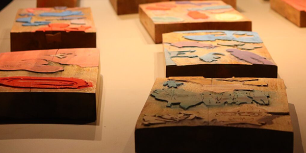 Na parte superior da imagem, aparece uma mesa expositiva de cor clara com diversos blocos retangulares de madeira distribuídos em fileiras. Sobre cada bloco há gravuras entalhadas e pintadas em diferentes cores, como azul, vermelho, rosa e bege, representando formas orgânicas e figuras estilizadas. Em primeiro plano, um dos blocos ocupa o centro da imagem, enquanto os demais se estendem em direção ao fundo, criando sensação de profundidade. A iluminação destaca as texturas da madeira e das gravuras, enquanto o ambiente ao redor permanece desfocado, concentrando a atenção nas peças expostas.