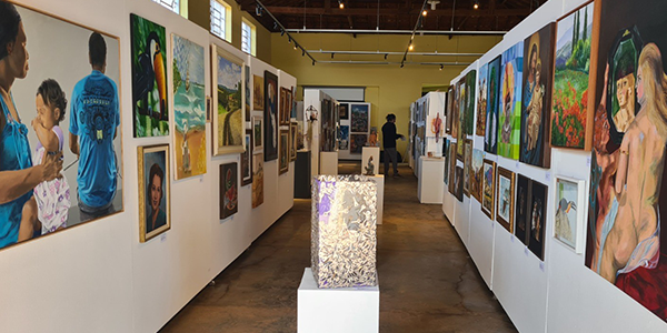 Na parte superior da imagem, aparece o interior de uma galeria de exposições com teto de madeira aparente e iluminação por trilhos. Ao centro, um corredor conduz o olhar até o fundo do espaço, onde há um visitante em pé. Nas laterais, painéis brancos exibem dezenas de pinturas de diferentes formatos, retratando paisagens, pessoas, cenas do cotidiano e figuras humanas. Em primeiro plano, sobre um pedestal branco, está uma escultura em tons claros, iluminada internamente. O ambiente é amplo, com piso de concreto, com entrada de luz natural.