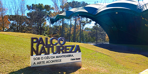 Na parte superior da imagem, vê-se uma área gramada sob céu azul. Em primeiro plano, à esquerda, há um letreiro instalado sobre uma base branca com a inscrição “PALCO NATUREZA” em letras grandes. Abaixo, lê-se: “SOB O CÉU DA MANTIQUEIRA, A ARTE ACONTECE”. Ao fundo, à direita, está o palco ao ar livre, coberto por uma estrutura metálica de formato curvo e cor verde-escura, sustentada por pilares finos. A área do palco encontra-se vazia. Ao redor, aparecem árvores de diferentes portes, algumas sem folhas, compondo a paisagem natural do Museu e Auditório.