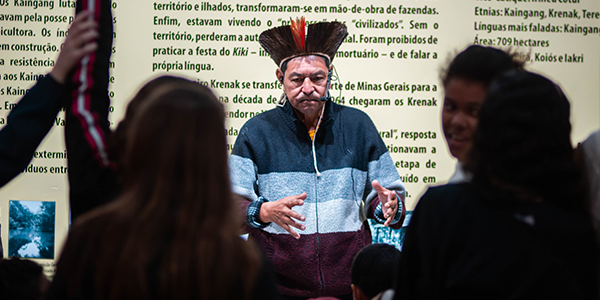 Na parte superior da imagem, aparece o interior de uma sala expositiva com uma parede em tom claro coberta por textos informativos em português. Ao centro, um homem indígena está de pé, voltado para o público. Ele usa um adereço de cabeça com penas em tons de marrom, vermelho e branco e veste um casaco de mangas longas nas cores azul-marinho, cinza e vinho. Com as mãos erguidas à frente do corpo, parece explicar algum tema aos visitantes. Em primeiro plano, diversas pessoas aparecem desfocadas, observando a apresentação; à esquerda, uma delas segura um celular na posição vertical, aparentemente registrando o momento. O ambiente é iluminado e conta com fotografias e painéis expositivos ao fundo.