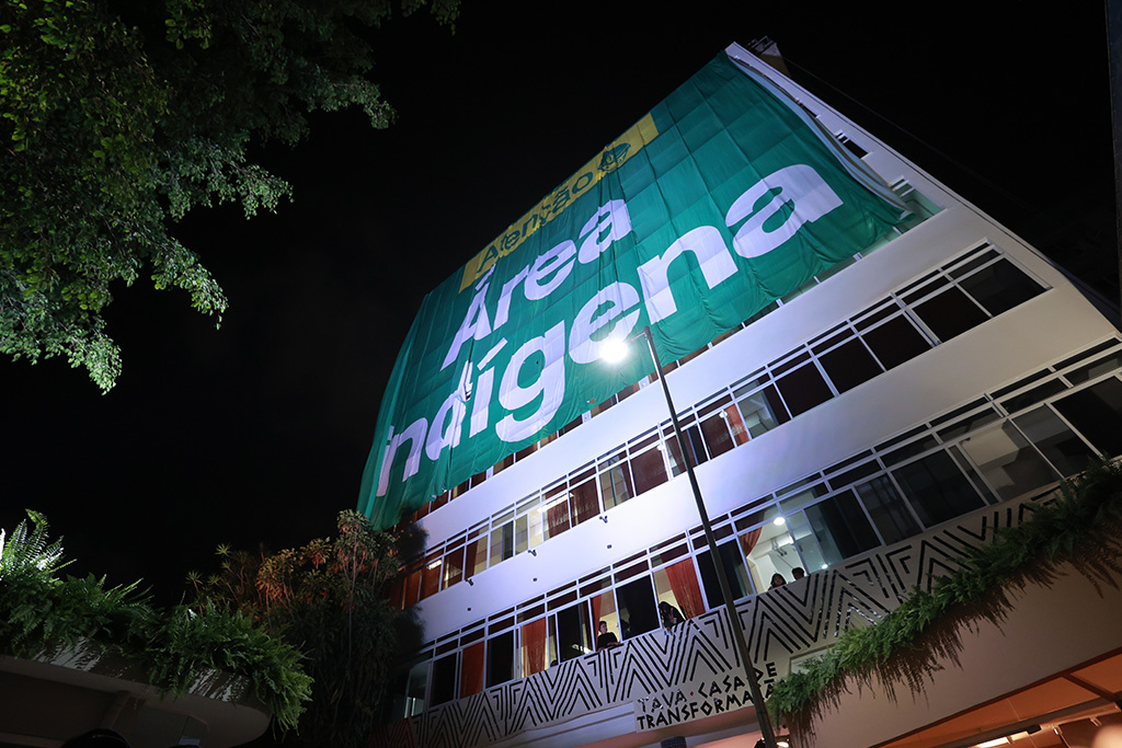 Fachada do museu, em imagem noturna. A foto mostra um prédio com um tecido sobreposto escrito "area indigena"