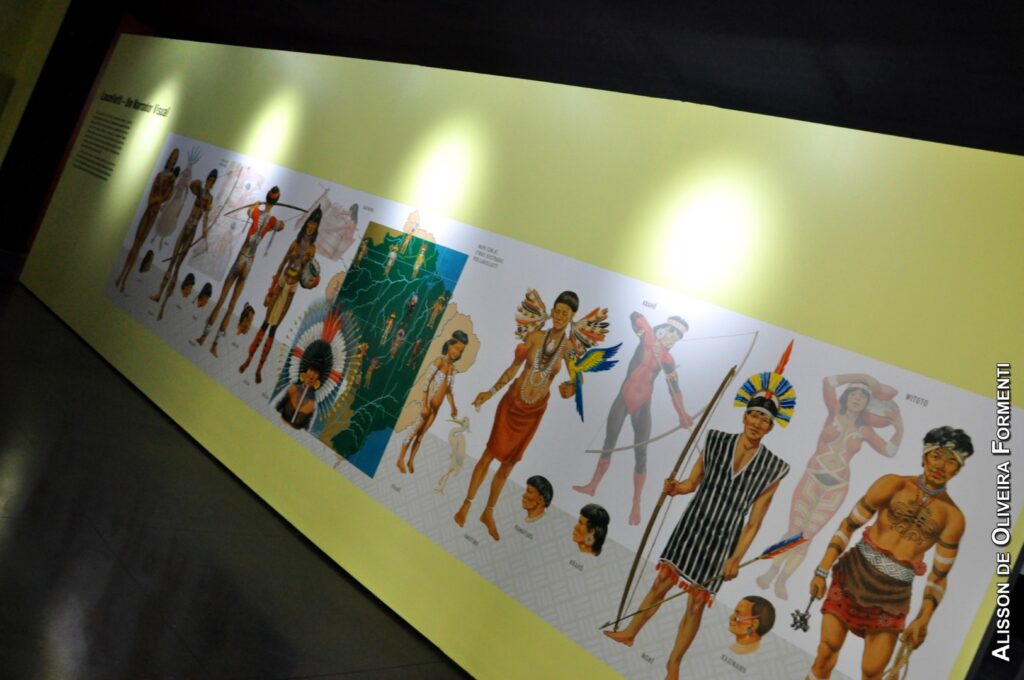 PraTodosVerem – A imagem mostra um painel amarelo com um cartaz branco contendo ilustrações de indígenas e seus adereços, como chapéus, arcos e flechas.   