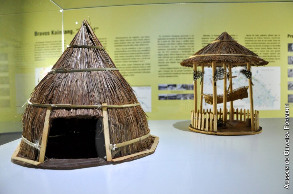 PraTodosVerem – A imagem mostra duas miniaturas arquitetônicas indígenas. Uma oca e um coreto com uma pequena rede de palha no centro. Eles estão sobre uma mesa branca, em frente a um painel amarelo com informações.