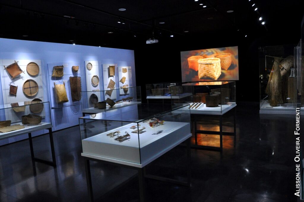 PraTodosVerem – A imagem mostra um saguão com chão escuro e redomas de vidros com objetos indígenas. A parede possui peças produzidas com materiais naturais como cestos e peneiras. 