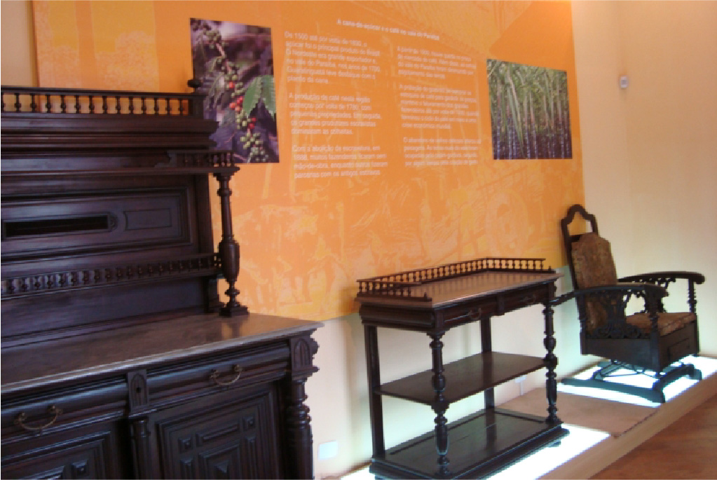 PraTodosVerem – A imagem mostra uma parede laranja com informações em branco e duas fotos de plantas. Na frente, três móveis antigos estão expostos sob uma plataforma luminosa: uma cadeira de balanço, um aparador e uma estante. Todos de madeira escura repletos de detalhes talhados. 