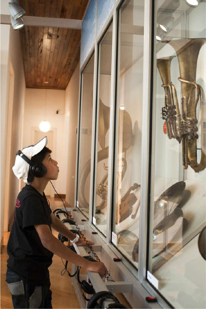 PraTodosVerem – A imagem mostra um menino com calça jeans, camiseta preta e boné branco com um fone nos ouvidos enquanto admira instrumentos musicais expostos na parede. 
