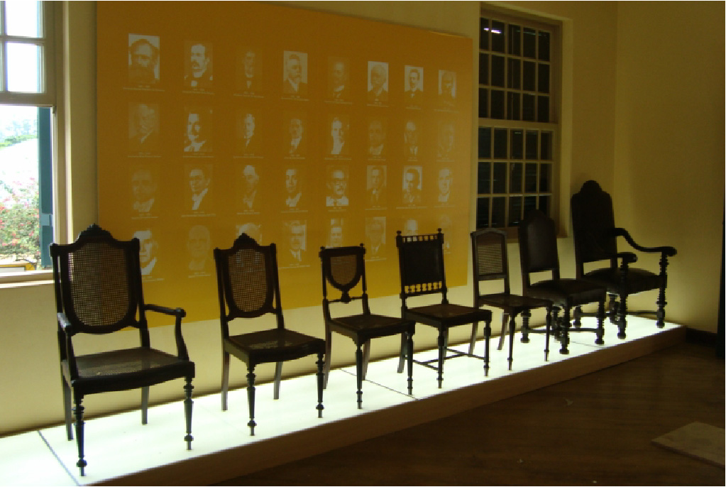PraTodosVerem – A imagem mostra sete cadeiras com detalhes expostas em uma plataforma baixa e luminosa. Atrás delas há um painel amarelo no centro de duas janelas com vitrais.