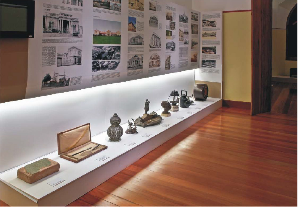 PraTodosVerem – A imagem mostra uma sala de museu com fotografias exibidas na parede e objetos expostos sob uma plataforma branca. O chão é de madeira e as paredes amarelas e brancas. 