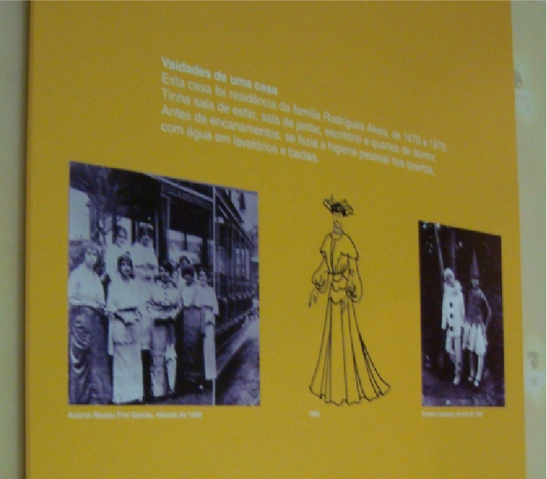 PraTodosVerem – A imagem mostra uma placa amarela contendo informações. A lateral esquerda exibe a foto de um grupo de mulheres, ao centro há um desenho de um croqui de moda da época, e na lateral direita tem duas pessoas vestidas de palhaços. 