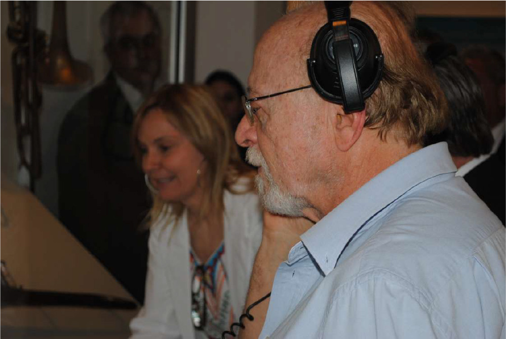 PraTodosVerem – A imagem mostra um homem de óculos, camisa social azul e fone de ouvido preto ao lado de uma mulher loira que está sorrindo. O fundo mostra outras pessoas próximas.