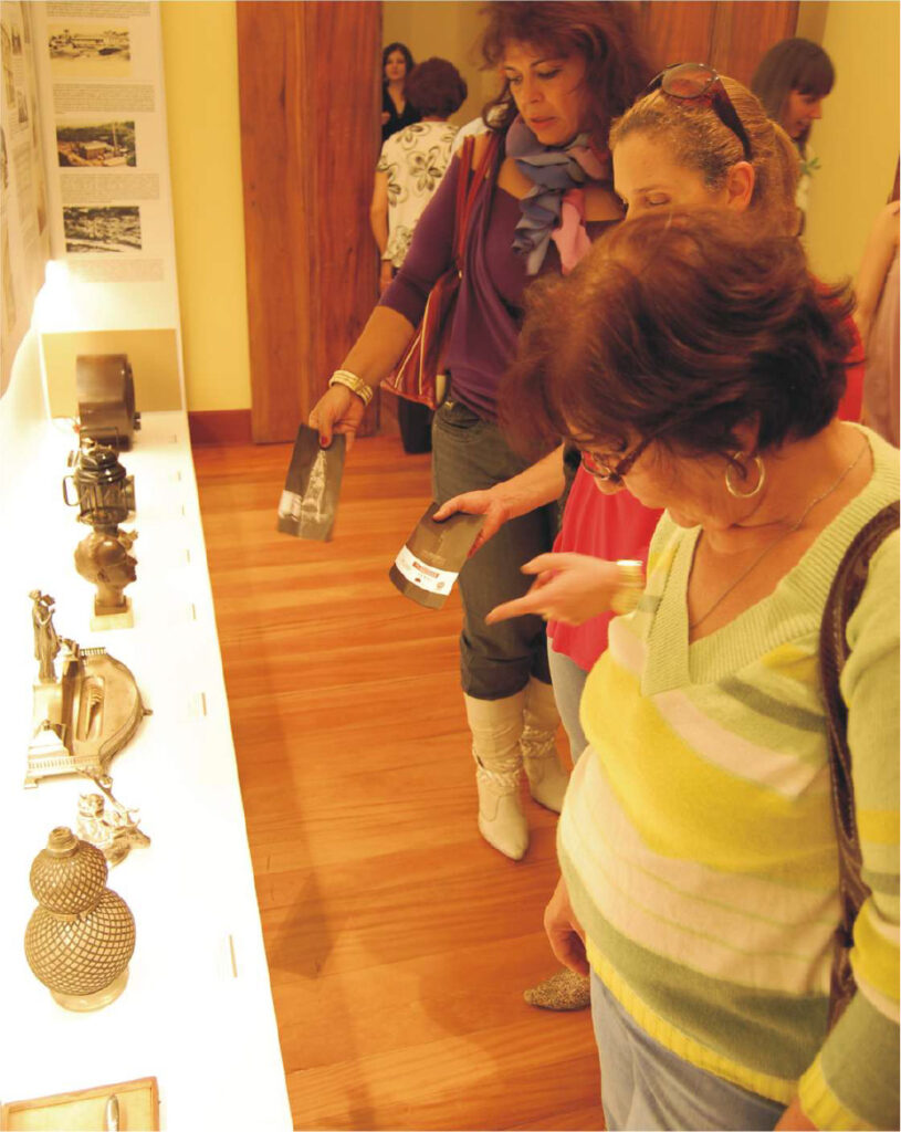 PraTodosVerem – A imagem retrata mulheres admirando peças de decorações antigas expostas em um balcão de museu.