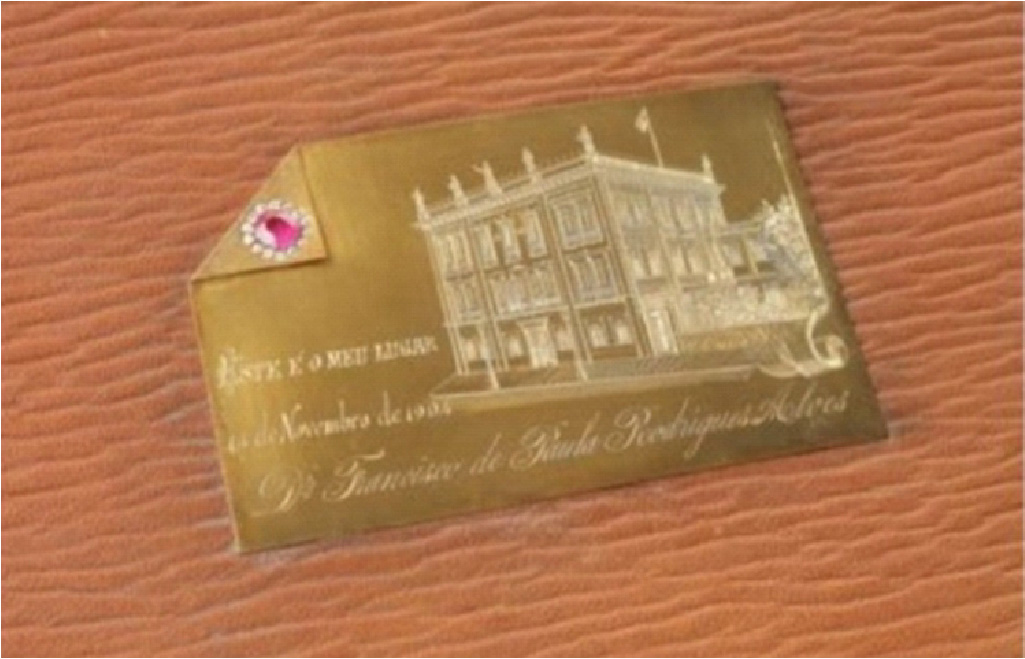 PraTodosVerem – A imagem mostra uma placa dourada com detalhes em branco. Uma casa antiga e frases gravadas com caligrafia cursiva. No topo da placa, uma pequena dobra no metal e uma pedra preciosa vermelha. 