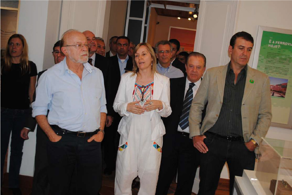 PraTodosVerem – A imagem mostra cerca de quinze pessoas reunidas caminhando pelas dependências do museu. A maioria homens, vestidos com roupas sociais. Uma mulher loira no centro da foto está vestida com uma roupa toda branca e detalhes coloridos. 