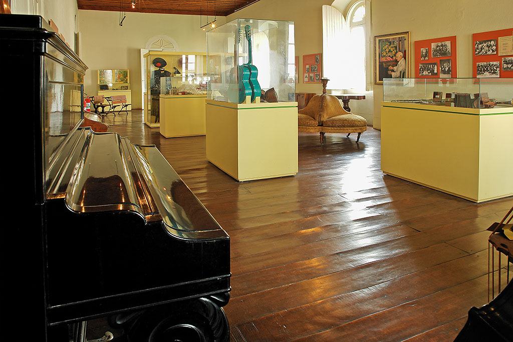 PraTodosVerem – Sala com chão de madeira e um piano marrom no canto esquerdo da imagem. Outros instrumentos musicais são expostos em redomas de vidro retangulares. As paredes são amarelas e a janela branca.  