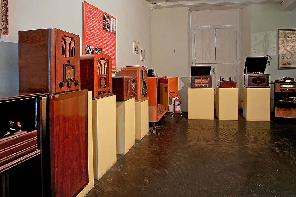 PraTodosVerem – A imagem mostra uma sala com aparelhos musicais antigos. Rádios e vitrolas de madeira são expostos em balcões amarelos. As paredes são brancas e na lateral esquerda há um painel vermelho com informações e fotografias em preto e branco.  