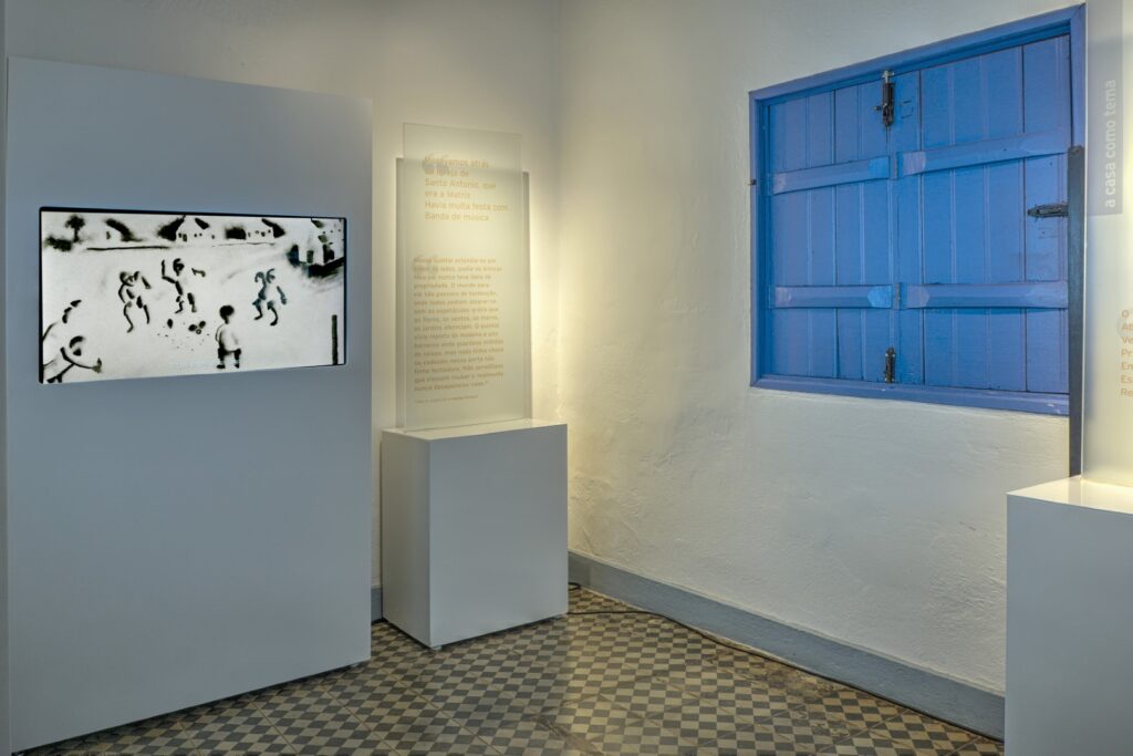 PraTodosVerem – A imagem mostra o interior de uma sala de museu. O chão é desenhado com pequenos ladrilhos em preto e branco. As paredes são brancas e a janela azul. Uma figura em preto e branco é exibida em um totem ao lado de um texto.  