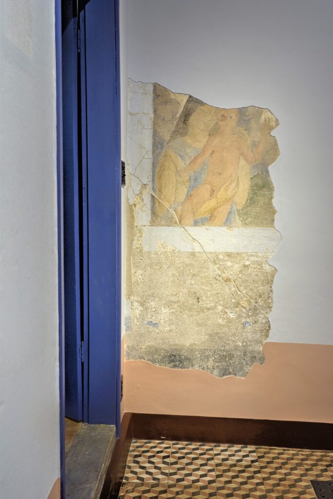 PraTodosVerem – A imagem mostra o batente de uma porta azul. No chão, uma soleira cinza com um rodapé de madeira e piso laranja e amarelo quadriculado. A parede é destacada por um restauro nas camadas mais antigas de tinta, mostrando a pintura de uma mulher segurando um bebê no colo.  