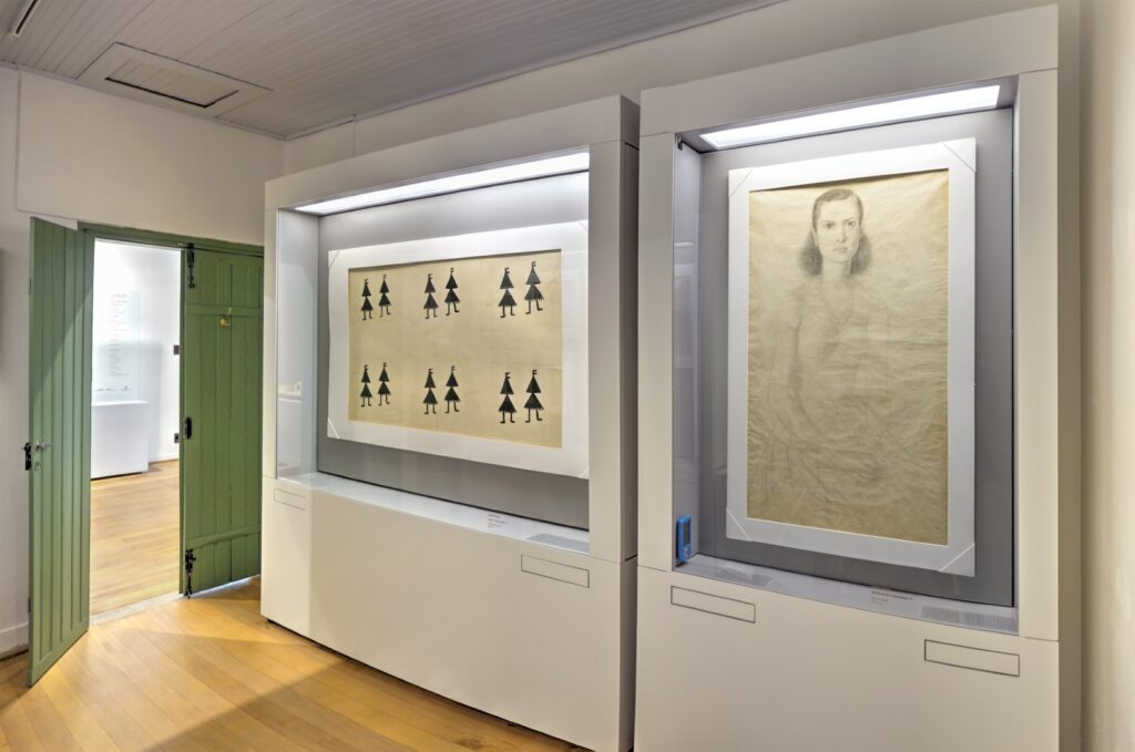 ParaTodosVerem: Uma sala com chão de madeira ripada na diagonal. Uma porta verde dupla com uma das folhas abertas. Na parede, duas vitrines que exibem obras de arte. 