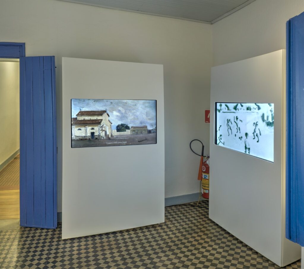 ParaTodosVerem: Uma sala com paredes brancas, portas azuis e chão quadriculado. Nas laterais, dois totens brancos suportam televisores.
